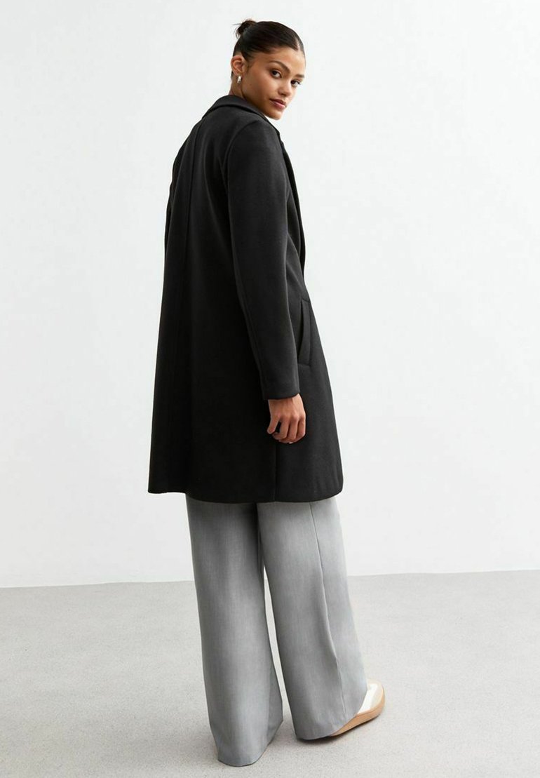 Manteau noir long avec un col cranté, des poches latérales et une finition texturée, associé à un pantalon large gris clair et des chaussures couleur crème.