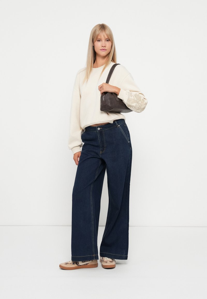 Szerokie ciemnoniebieskie dżinsy, kremowy sweter z ozdobnymi rękawami i szara torebka. Obuwie z kwiecistym wzorem. Proste tło.