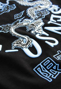 Svart tyg med en blå och vit drakegrafik, svirls av molnmönster och framträdande stiliserad text. Platt struktur med detaljerad design.