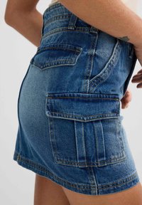 Falda mini de mezclilla en azul medio, con un diseño ajustado, bolsillos laterales y costuras visibles. Textura suave con ligero desgaste.