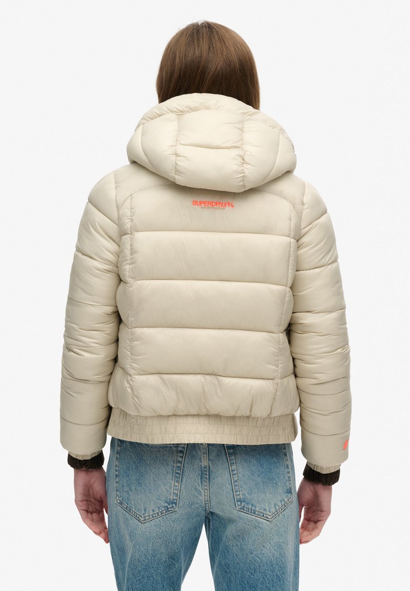 Beige Steppjacke mit Kapuze, mit gestepptem Design, elastischen Bündchen und einem orangefarbenen Logo auf dem Rücken. Kombiniert mit blauen Jeans.