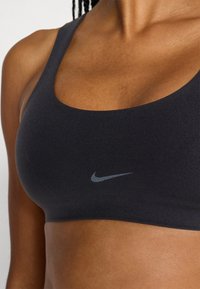 Μαύρο αθλητικό μπουστάκι από λείο ύφασμα με ντεκολτέ σε σχήμα λαιμόκοψης. Διαθέτει διακριτικό λογότυπο Nike σε γκρι στο μπροστινό μέρος.