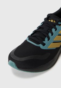 Baskets de sport noires avec des accents turquoise, dotées d'un upper en maille et d'une semelle texturée. Des rayures dorées ajoutent des détails, et les boucles de lacets garantissent un maintien sécurisé.