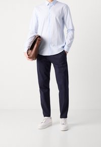 Camisa de botones azul claro, pantalones azul marino, zapatillas blancas, sosteniendo un bolso de mensajero de cuero marrón. Diseño casual y moderno con elementos clásicos.