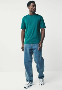 Zöld pamut póló rövid ujjakkal, kék farmernadrággal párosítva. A pólón kerek nyakkivágás található, és szabványos slim fit fazonú.