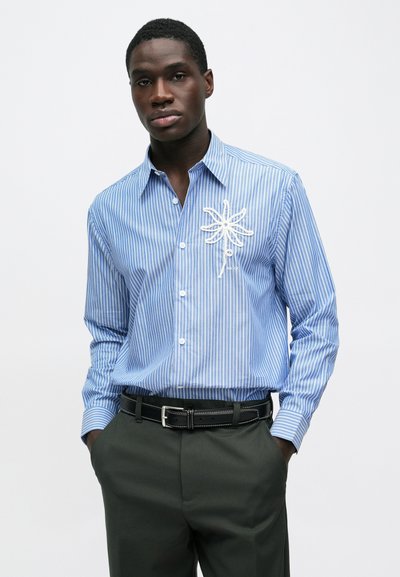 Chemise boutonnée à rayures bleues et blanches avec un motif brodé d'une araignée blanche sur la poitrine, associée à un pantalon vert foncé et une ceinture noire.