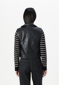 Person mit glattem schwarzem Haar trägt eine schwarze, kurze Lederweste über einem schwarz-weiß gestreiften Langarmshirt und dunklen Jeans, Rückansicht.