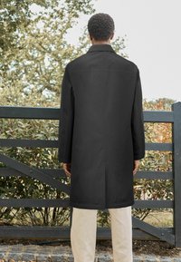 Manteau noir à coupe droite, avec un col et des manches longues, porté par-dessus un pantalon de couleur claire, sur un fond vert.