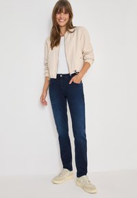 Chaqueta bomber beige con puños elásticos, camiseta blanca, jeans azul oscuro y zapatillas de color claro, con un diseño simple y corte ajustado.