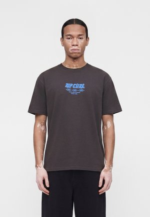 Vitiligós férfi fekete Rip Curl pólóban és fekete nadrágban, tiszta fehér háttér előtt, előre nézve.