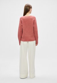 Roza pletena jopica z sproščenim krojem, ki ima okroglo izrez in izdelavo z gumbom na hrbtni strani, v kombinaciji s kremnimi širokimi hlačami.