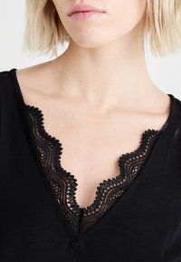 Μαύρη μπλούζα με V-λαιμόκοψη που διαθέτει γυαλιστερή μαύρη δαντέλα με φίνιρισμα σε σχήμα κυματοειδούς. Λείο βαμβακερό ύφασμα με άνετη εφαρμογή γύρω από το λαιμό και τους ώμους.