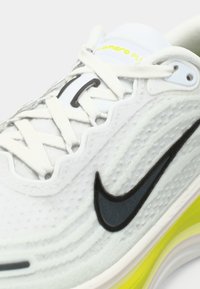 Κοντινή λήψη λευκού αθλητικού παπουτσιού Nike με μαύρο λογότυπο, υφαντό επάνω μέρος, λευκές κορδόνες και κίτρινη λεπτομέρεια στα μαξιλαράκια της σόλας.