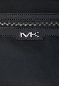 Borsa a tracolla in nylon nero con zip argentata, caratterizzata da una patch in pelle texturizzata con il logo "MK" centrato sopra la zip. Finitura liscia complessiva.
