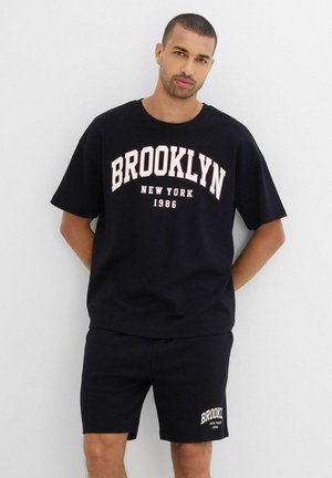 Set di t-shirt e pantaloni corti in cotone nero con grafica "BROOKLYN NEW YORK 1986" in bianco e rosso. Collo arrotondato, maniche corte, vestibilità rilassata.