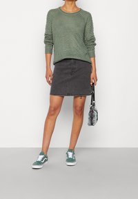 Olivevihreä neulepaita, jossa on teksturoitu kuvio, harmaat farkku-mini-hame ja turkoosit tennarit. Musta-valkoraidallinen laukku pidetään kädessä.