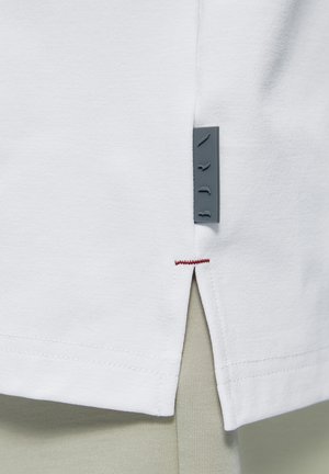 Biely okraj odevu s bočným rázporkom, červeným prešitím na vrchu rázporku a malou sivou štítkou z látky pripevnenou blízko rázporku.