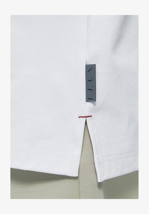 Biely okraj odevu s bočným rázporkom, červeným prešitím na vrchu rázporku a malou sivou štítkou z látky pripevnenou blízko rázporku.