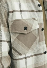 Lähikuva vaaleanbeige ja valkoinen ruudullinen takin tasku mustalla napilla, jossa on suuri läppä ja kontrastinen vino kuvio.