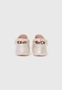 Crocs rose clair à finition brillante, avec des brides blanches décoratives ornées de petites perles. Le talon affiche le logo.