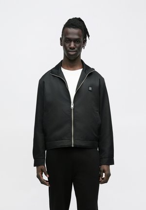 Hombre joven con cabello oscuro trenzado que lleva una chaqueta negra con cremallera sobre una camiseta blanca y pantalones negros, de pie frente a un fondo liso.