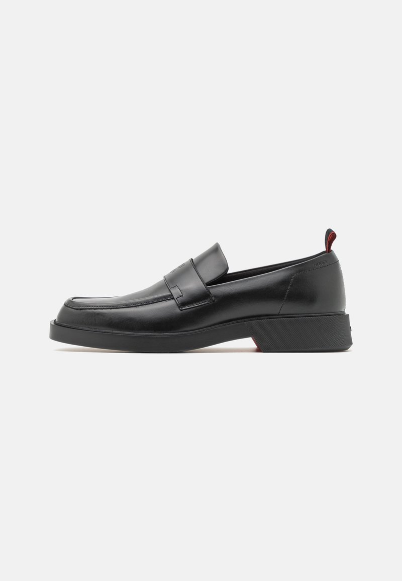 Mocassin noir en cuir à enfiler avec un petit talon, bout carré arrondi et languette en tissu rouge à l'arrière, présenté sur fond blanc.