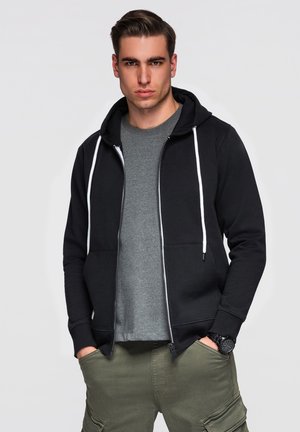 Hombre joven vistiendo una sudadera negra con cremallera y cordones blancos, camiseta gris, pantalones cargo oliva, manos en los bolsillos y reloj de pulsera negro.