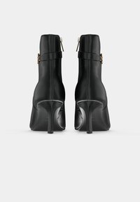 Stivaletti neri in pelle con tacco a stiletto alto, zip posteriore e una fascia con un accento dorato alla caviglia. Texture liscia, design elegante.