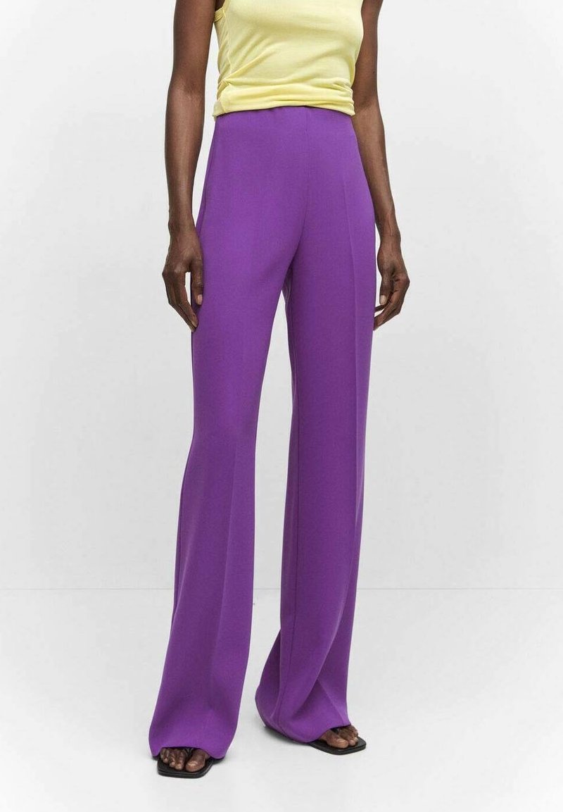 Personne portant un pantalon large violet brillant, un haut sans manches jaune et des sandales noires à bouts ouverts, debout devant un fond blanc uni.