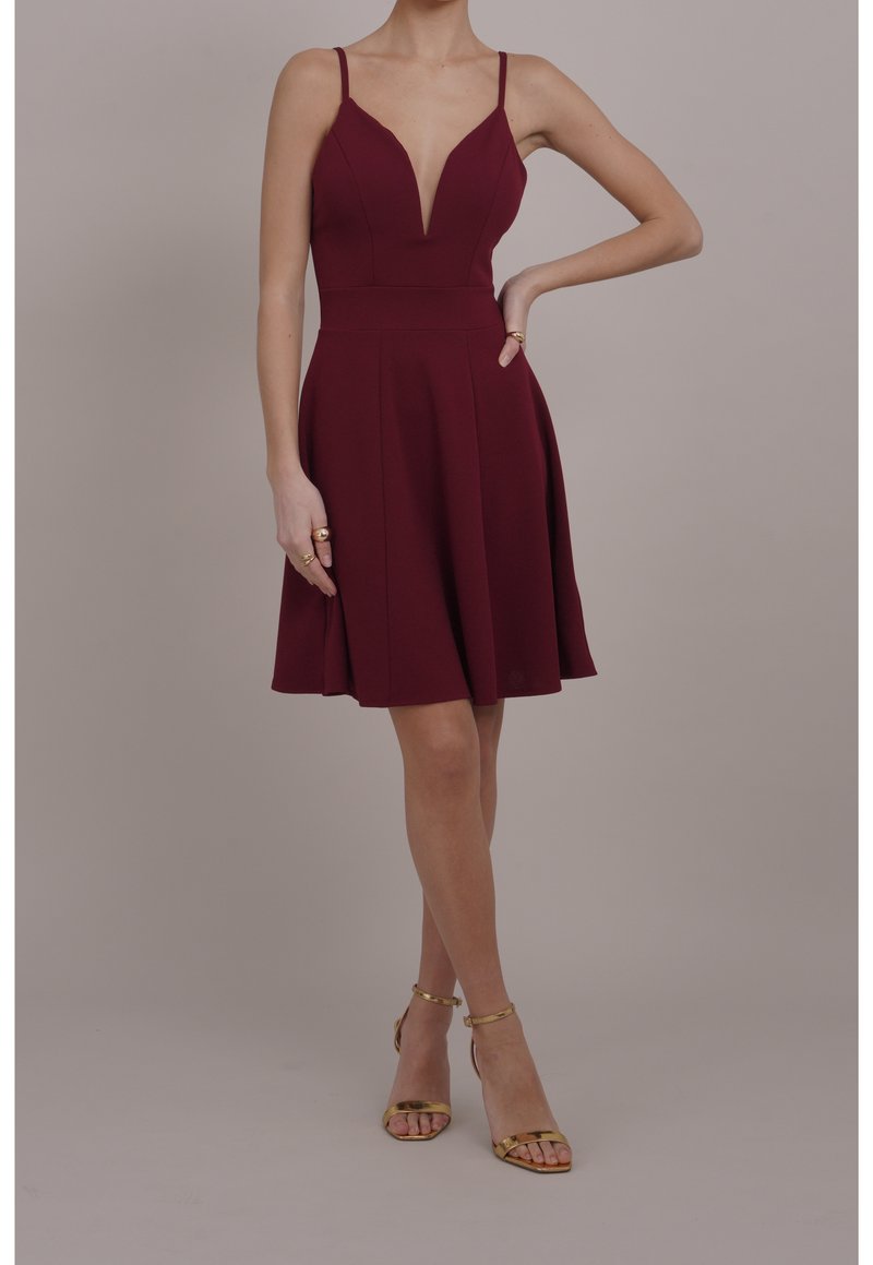 Femme portant une robe bordeaux sans manches avec un profond décolleté en V, jupe évasée et sandales dorées à talons hauts avec bride à la cheville, debout avec une main sur la hanche.