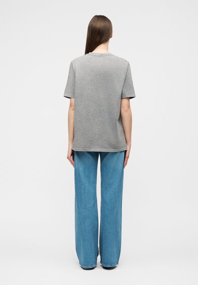 Άτομο με μακριά καστανά μαλλιά στέκεται γυρνισμένο πίσω, φορώντας μια ξεθωριασμένη γκρι κοντομάνικη μπλούζα και άνετο μπλε τζιν, σε μονόχρωμο φόντο.