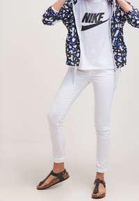 Vita smala byxor i kombination med en svart och blåmönstrad jacka samt en vit t-shirt med Nike-logotyp. Skorna är svarta platta sandaler med korksula.