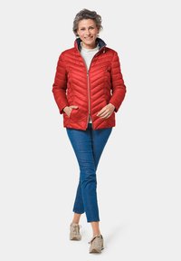 Veste matelassée rouge avec poches latérales, fermeture éclair avant et col intérieur bleu marine, associée à un jean bleu et des baskets beiges.