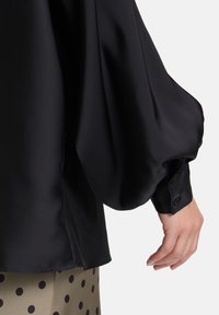 Blusa nera in raso con maniche voluminose e polsini abbottonati, caratterizzata da una texture liscia e da una leggera lucentezza. Il design notevole include un tessuto fluente.