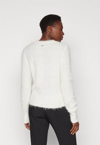 Maglione bianco peloso con maniche lunghe, scollo a girocollo e orlo a coste. Presenta un piccolo dettaglio del logo metallic al centro del retro dello scollo.