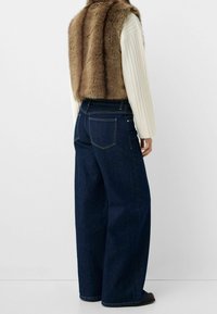 Brown faux fur vest over a ribbed white long-sleeve top, paired with wide-leg dark blue denim jeans, featuring back pockets and visible stitching.