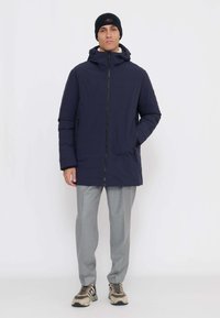 Giacca imbottita blu navy con cappuccio, zip frontale e tasche laterali, abbinata a pantaloni grigio chiaro e sneaker beige. Berretto nero in testa.