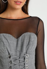 Femme portant des boucles d'oreilles créoles et un haut noir transparent sous un corset gris à carreaux avec un laçage devant.