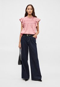 Roza bluzica s volanima i smock detaljima, uparena s tamnim širokim trapericama i crnom torbicom.