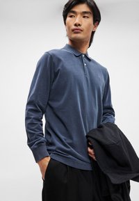 Ναυτική μακρυμάνικη πόλο μπλούζα με τριπλή κουμπότρυπα, ριμπ μανσέτες και διακριτική λεπτομέρεια λογότυπου, συνδυασμένη με μια μαύρη υφασμάτινη ζακέτα.