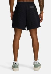Man som bär svarta, texturerade sportbyxor med en bakficka med dragkedja, vita Under Armour-strumpor och ljusgröna sneakers, stående vänd bort.