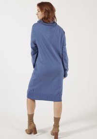 Robe en tricot bleue avec un col montant, des manches longues et un ourlet côtelé. Portée avec des bottines marron à la cheville. Tissu texturé.