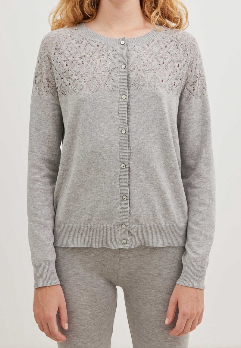 Femme portant un cardigan boutonné gris clair avec un motif en maille dentelle sur la partie supérieure de la poitrine et un pantalon gris assorti.