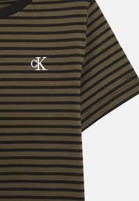 Olivgrünes und schwarzes gestreiftes T-Shirt mit Rundhalsausschnitt, das ein weißes besticktes "ck"-Logo auf der Brust und den Schriftzug "CALVIN KLEIN" auf dem Ärmel trägt.