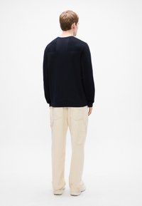 Persoană întoarsă cu spatele, îmbrăcată într-un tricou lung negru, pantaloni cargo bej și pantofi albi, pe un fundal alb simplu.