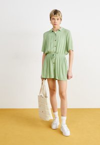 Combinaison verte à manches courtes avec des boutons, taille élastique et cordon de serrage. Associée à un sac fourre-tout beige et des baskets plateformes blanches.