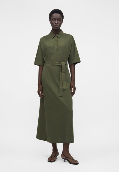 Ψηλή γυναίκα που φοράει μεσάτο ελιάς πράσινο φόρεμα με ζώνη, γιακά, κοντά μανίκια, κουμπιά και μαύρα σανδάλια με λουράκια και τακούνι.