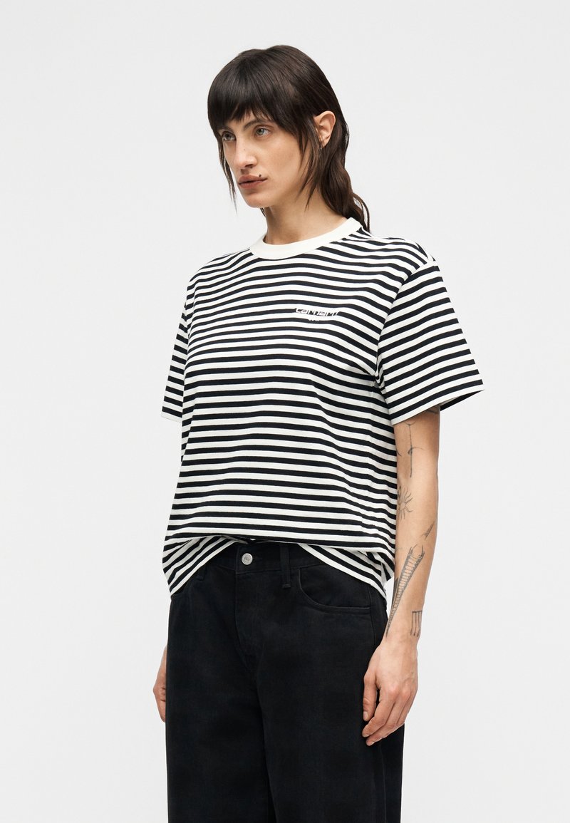 Maglietta a righe bianche e nere con collo rotondo e maniche corte. Presenta un piccolo logo sul petto. Abbinata a pantaloni neri.