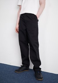 Black cotton pants with a relaxed fit, featuring side pockets and an elastic waistband, paired with black sneakers on a blue carpet.