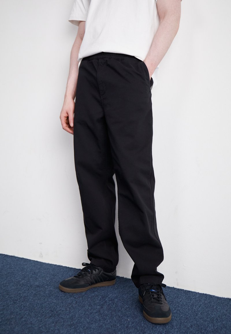 Black cotton pants with a relaxed fit, featuring side pockets and an elastic waistband, paired with black sneakers on a blue carpet.